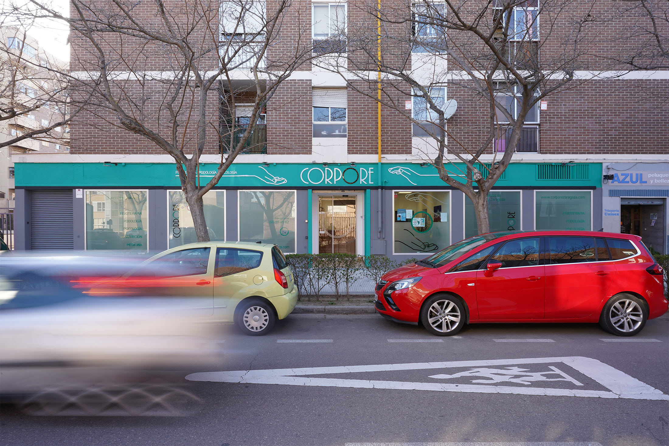 Fachada exterior a pie de calle de la clínica con cerramientos en color turquesa bajo un edificio de ladrillo